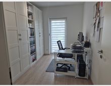 Schmales, helles Homeoffice mit hellem Holzfußboden, großem weißen Einbauschrank und Regalen mit Ordnern an der linken Wand. Rechts steht ein weißer Schreibtisch mit Computer und Ablagen vor einem Fenster mit Doppelrollo; an der Wand hängt ein Pinnbrett mit Zetteln.