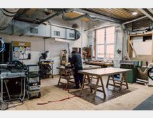 Werkstatt mit Holzboden, ausgestattet mit Werkbänken, Holzverarbeitungsmaschinen, Werkzeugwänden und einem großen Fenster, das natürliches Licht hereinlässt. Eine Person arbeitet an einem Werkstück in der Mitte des Raums.