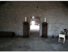 Ein leerer Raum mit grauer, unverputzter Steinwand und einer Türöffnung in der Mitte, flankiert von zwei kleinen Säulen mit weißen Kerzen darauf. Im angrenzenden Raum sind graffitibeschmierte Wände, alte Möbel und gelagerte Gegenstände sichtbar.