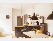 Helle Küche mit skandinavischem Design, bestehend aus einer Arbeitsfläche mit schwarzer Wandtafel, auf die "Hello" geschrieben ist, einer rustikalen Holzoberfläche, Hängelampen, einem Kaminofen, einer Kaffeemaschine und offenen Regalen. Im Vordergrund steht ein Holztisch mit Blumen in einer Vase.