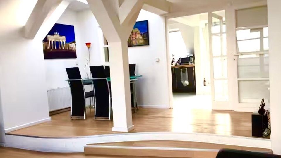 Offener Wohn- und Essbereich mit hellem Holzfußboden, sichtbaren weißen Dachbalken und einer leicht erhöhten Stufe zum Essplatz mit Glastisch und schwarzen Stühlen. Im Hintergrund führen Glastüren in einen angrenzenden Raum; an den Wänden hängen gerahmte Bilder.