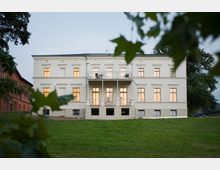 Großes, weißes, zweistöckiges Herrenhaus im klassischen Stil mit einer zentralen, erhöhten Terrasse und vielen gleichmäßig angeordneten Fenstern, umgeben von einer grünen Rasenfläche und vereinzelten Bäumen. Im Hintergrund ist der Himmel leicht bewölkt.