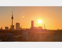 Silhouette der Skyline von Berlin bei Sonnenuntergang mit dem Fernsehturm als markantem Element. Im Vordergrund sind Dächer und Geländer sichtbar, während die Sonne den Himmel in orangefarbenes Licht taucht.