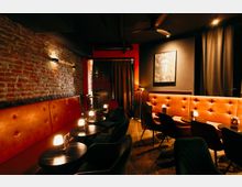 Innenraum einer gedimmten Bar-Lounge mit freiliegender Backsteinwand, gepolsterten orangefarbenen Sitzbänken und kleinen runden Tischen mit Tischlampen. Dunkle Decke mit Lüftungsrohren und Ventilator, schwarze Vorhänge sowie ein gerahmtes Bild an der Wand.