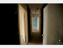 Ein dunkler, schmaler Flur in einem verlassenen Gebäude mit beschädigten Wänden und sichtbaren Kabeln an der Decke. Mehrere offene Türen führen in andere Räume, wobei eine Wand in der Ferne blau gestrichen ist.