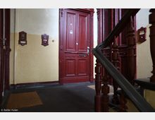 Treppenhaus eines Wohnhauses mit roten Holzdetails, darunter eine dekorative Tür mit Zierleisten und ein geschnitztes Treppengeländer aus dunklem Holz. Die Wände sind beige gestrichen und mit roten Klingelschildern versehen.