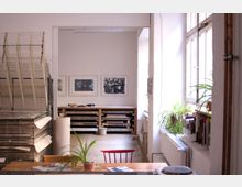 Heller Raum eines Ateliers mit großen Fenstern auf der rechten Seite, Pflanzen auf Fensterbänken, einem Holztisch mit roten Stuhl im Vordergrund und einem Trockenregal sowie Kunstwerken an der Wand im Hintergrund.