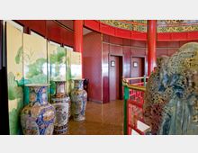 Innenraum mit asiatischer Dekoration, einschließlich großer bemalter Porzellanvasen, eines Paravents mit floralen Motiven und roten Holzwänden mit Ornamenten. Im oberen Bereich ist eine bunte, verzierte Decke mit traditionellen Mustern sichtbar.