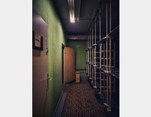 Schmaler Innenflur mit grün gestrichenen Wänden, Neon-Deckenleuchte und gemustertem Bodenbelag. Rechts stehen hohe, metallene Rohrrahmen-Regale, links befinden sich Türen und Schaltschränke; am Ende steht ein kleiner Wagen.