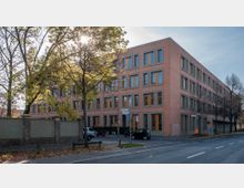 Backsteingebäude mit moderner Fassade und großen rechteckigen Fenstern, gelegen an einer Straßenecke mit Straßenschildern und Fußwegen, umgeben von Bäumen, die teilweise belaubt sind. Im Vordergrund sind eine Mauer, ein kleiner Parkplatz mit Autos und ein Motorrad sichtbar.