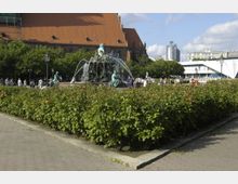 Ein Park mit gepflasterten Wegen und einem zentralen Brunnen, der Figuren sowie Wasserspritzer zeigt. Im Hintergrund sind ein Gebäude mit einem roten Dach und moderne Gebäude sichtbar, umgeben von Bäumen und Grünflächen.