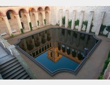 Innenhof einer historischen Steinmaueranlage mit einem rechteckigen Wasserbecken in der Mitte, das die umliegenden Bögen und Wände spiegelt. Die Umgebung ist mit Kletterpflanzen bewachsen, und Sitzbänke stehen entlang der Wände.