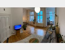 Wohnzimmer einer Altbauwohnung mit Holzdielenboden, großen Sprossenfenstern und einem grauen Sofa. Im Raum stehen mehrere Zimmerpflanzen, ein kleiner Fernseher, ein runder Couchtisch und ein Esstisch mit Stühlen; im Hintergrund ist ein Schreibtisch vor einem Fenster zu sehen.