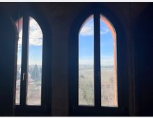 Zwei gotische Spitzbogenfenster mit schwarzem Rahmen geben den Blick auf eine ländliche Landschaft frei, bestehend aus Feldern, Bäumen und einem kleinen Dorf unter einem sonnigen Himmel mit vereinzelten Wolken. Ein roter Ziegelrahmen ist bei einem der Fenster sichtbar.