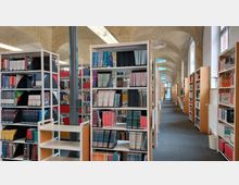 Innenansicht einer Bibliothek mit hohen Bücherregalen, die mit Büchern und Zeitschriften gefüllt sind. Das Gebäude hat gewölbte Decken aus gelbem Backstein, große Fenster auf der rechten Seite und einen langen Teppichboden-Gang in der Mitte.