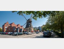 Eine historische Windmühle mit dunklem Holzdach und Flügeln steht zentral in einem städtischen Umfeld, umgeben von Backsteingebäuden, parkenden Autos und Bäumen. Im Hintergrund sind moderne Wohnhäuser sichtbar.