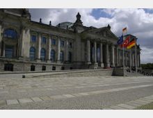 Historisches Gebäude mit Säulenfassade und großen Fenstern, erkennbar als der Reichstag in Berlin; im Vordergrund eine gepflasterte Terrasse, und mehrere Fahnen, darunter die deutsche, europäische und eine türkische Flagge, wehen im Wind. Im Hintergrund ist eine Glaskuppel zu sehen.