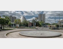 Städtischer Platz mit einem leeren, runden Brunnen im Vordergrund, umgeben von Gehwegen und Bäumen. Im Hintergrund stehen moderne Hochhäuser, eine futuristisch geformte Kuppel und eine U-Bahn-Station, daneben Verkehrswege mit Autos, Fußgängern und Ampeln.