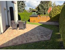 Gepflasterte Terrasse mit einem Gartentisch und sechs Stühlen aus Rattan, angrenzend an eine weiße Hauswand. Im Hintergrund ein Holzgerätehaus, grüne Hecken, Bäume und ein Rasenbereich.