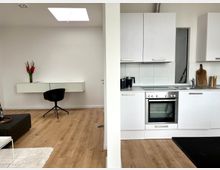 Helles, modernes Wohnraum mit offener Küche und minimalistischer Einrichtung. Linke Seite: ein schwarzer Stuhl vor einem weißen, schwebenden Schreibtisch; rechte Seite: weiße Küchenzeile mit Ofen, Spüle und Hängeschränken, dazu helle Holzfußböden und Tageslicht.