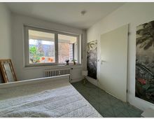 Schlafzimmer mit weiß gestrichenen Wänden, einem großen Fenster mit Blick auf einen Balkon und eine Außenlandschaft. Im Raum befinden sich ein Bett mit heller Tagesdecke, Pflanzenmotiv-Tapeten an der Wand, ein Heizkörper unter dem Fenster, ein kleiner Nachttisch mit Lampe und ein schräg angelehnter Spiegel.