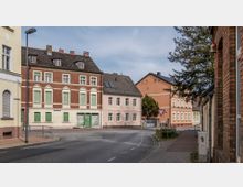 Straßenecke in einer Kleinstadt mit mehrstöckigen Wohnhäusern; links ein Gebäude mit rotem Ziegelmauerwerk und grünen Fensterläden, rechts weitere Häuser in Pastellfarben. Ein Gehweg verläuft entlang der Straße, die mit einem Schild eine Vorfahrtsregelung anzeigt.