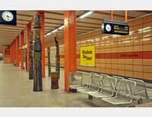 U-Bahnhof mit orangefarbenen Stahlstützen, beigefarbenen Wandfliesen und mehreren Holzskulpturen. Im Vordergrund befinden sich Sitzbänke aus Metall sowie eine Uhr und Anzeigetafel mit Zuginformationen.