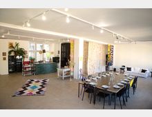 Ein heller, offener Raum mit Betonboden, einer langen gedeckten Tafel, einer Sitzecke mit weißen Sofas sowie einer Küchenzeile mit grünen Fliesen. Bunte Vorhänge und dekorative Elemente wie ein Teppich und Pflanzen ergänzen das moderne Design.