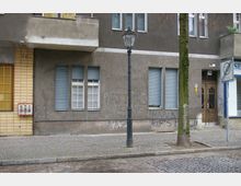 Wohnhausfassade in einer städtischen Umgebung mit grauer Putzfassade, blau geschlossenen Fensterläden und einer Holzeingangstür. Davor befindet sich ein Gehweg mit Kopfsteinpflaster, ein Laternenmast und ein Baum; an der Hauswand sind Graffiti sichtbar.