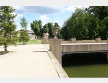 Ein kleiner Park mit einer steinernen Brücke über einem Wasserlauf, flankiert von dekorativen Steinvaselementen. Im Hintergrund sind Rasenflächen, Bäume und eine Mauer mit dahinterliegenden Gebäuden sichtbar.