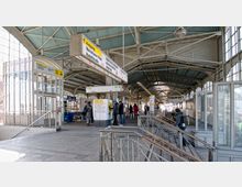 Heller Bahnhof mit überdachtem Bahnsteig und Glaswänden, auf dem mehrere Fahrgäste stehen. Zu sehen sind Fahrkartenautomaten, Wegweiser, ein Lift sowie Schienen und Zugangsbereiche zu den Gleisen.