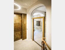 Innenansicht eines Treppenhausflurs mit hohen Rundbögen, weiß verputzten Wänden und einem modernen Ring-Deckenlicht. Links befindet sich eine messingfarbene Aufzugstür, rechts eine gläserne Tür mit goldenem Rahmen, die in einen Gang mit freigelegter Ziegelwand und Briefkastenanlage führt.