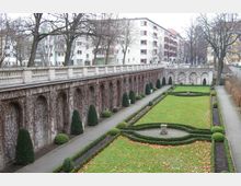 Formaler Garten mit symmetrischen Rasenflächen, umgeben von niedrigen Hecken und Wegen, flankiert von einer mit Kletterpflanzen bewachsenen Steinmauer mit Rundbögen. Im Hintergrund sind mehrstöckige Wohngebäude und kahle Bäume sichtbar.