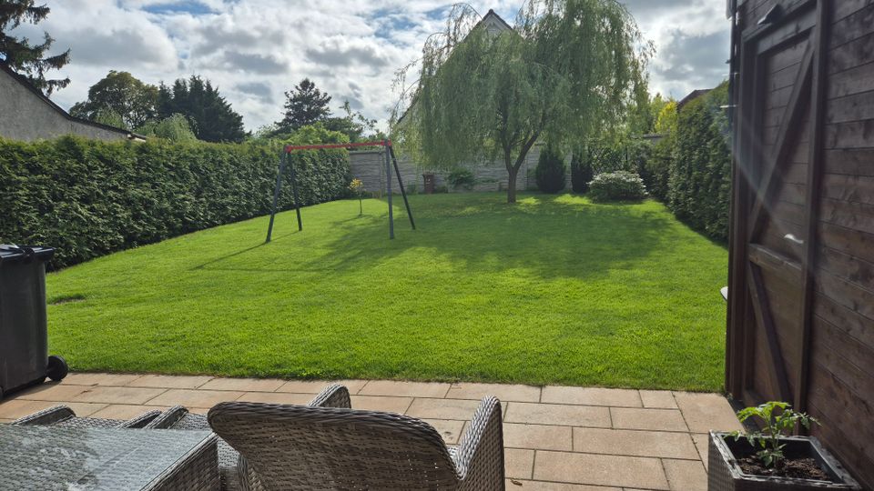 Blick von einer gepflasterten Terrasse mit Rattanmöbeln auf einen großen, gepflegten Rasen im eingezäunten Garten. In der Mitte stehen eine Schaukel und ein großer Baum, umgeben von hohen Hecken sowie einem Holzgartenhaus am rechten Bildrand.