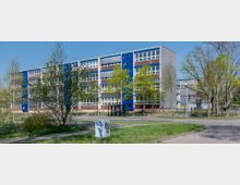 Ein vierstöckiges Schulgebäude mit roten und blauen Fassadenelementen sowie großen Fenstern, umgeben von Bäumen und einem Grünbereich. Im Vordergrund befinden sich ein Gehweg, Rasengestaltung und ein kleiner überdachter Spielbereich.