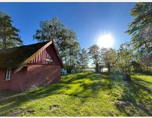 Kleines rotes Holzhaus mit steilem, moosbewachsenem Satteldach am Rand einer grünen Wiese, umgeben von hohen Bäumen. Im Hintergrund sind mehrere kleine weiße Häuschen aufgestellt, dahinter glitzert ein See unter tief stehender Sonne am klaren Himmel.