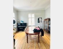 Esszimmer mit Holzdielenboden, einem länglichen Holztisch und transparenten Plastikstühlen. Im Hintergrund befindet sich ein schwarzes Klavier, ein Schrank mit Gläsern und Büchern sowie ein großes Fenster, durch das Tageslicht hereinfällt.