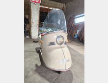 Innenraum einer Werkstatt oder Lagerhalle mit Betonboden, rauen Wänden und gestapelten Holzbrettern sowie Beleuchtungsständer im Hintergrund. Im Vordergrund steht ein kleines, beige lackiertes dreirädriges Kabinenfahrzeug mit großer gewölbter Windschutzscheibe und zentralem Rundscheinwerfer.