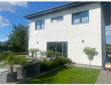 Zweistöckiges, modernes Wohnhaus mit weißer Fassade und großen Fenstern, gesehen vom Garten mit Rasenfläche. Im Vordergrund stehen eine Terrasse mit dunklen Loungemöbeln sowie Beete mit Ziergräsern und kleinen Palmen.