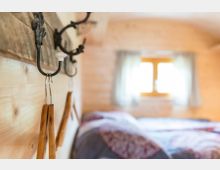 Ein gemütliches Schlafzimmer mit Holzwänden, einem Fenster mit Vorhängen im Hintergrund und einem Bett mit gemusterten Decken. Im Vordergrund sind Haken an einer Holzwand mit daran hängenden Holzgriffen zu sehen.