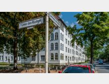 Straßenszene mit Wohngebäude in heller Putzfassade, hohen, bogenförmigen Fenstern und roten Ziegeldächern, flankiert von dicht belaubten Bäumen. Im Vordergrund steht ein Straßenschild „Unter den Rüstern“, unter anderem sind geparkte Autos und ein Bürgersteig zu sehen.