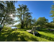 Wiese am Ufer eines Sees mit hohen Laubbäumen und Blick auf einen kleinen Steg im Wasser. Im Vordergrund stehen eine hölzerne Gartenliege und ein Metallgartentisch mit Stühlen, umgeben von Sträuchern und einzelnen Steinen unter blauem Himmel.