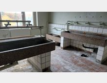 Ein verlassener Waschraum mit mehreren großen, grob gemauerten Waschbecken aus Stein. Die Wände sind mit weißen Keramikfliesen verkleidet, der Boden besteht aus abgenutzten, rötlichen Fliesen, und im Hintergrund befindet sich ein Fenster mit Blick ins Freie.