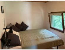 Kleines Schlafzimmer mit Doppelbett, dunklen Kissen und grünlicher Decke, flankiert von einer Stehlampe. Rechts ein Holzfenster mit teilweiser Jalousie und Blick auf dichtes grünes Laub draußen, dazu helle Wände und Holzdecke.