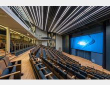 Ein moderner Hörsaal mit ansteigenden Sitzreihen aus Holz- und Polstermaterial und einem großen LED-Bildschirm an der Vorderwand. Die Decke ist mit parallelen LED-Leisten beleuchtet, und es gibt eine Galerieebene mit Glasgeländer auf der linken Seite.