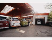 Eine offene Werkstatt mit einem überdachten Bereich, unter dem mehrere bunte VW-Busse aus der Retro-Ära geparkt stehen. Im Hintergrund ist eine weiße Ziegelwand mit einer offenen Garagentür zu sehen, in der sich ein weiterer VW-Bus befindet.