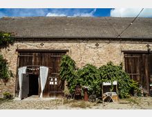 Eine alte Backstein-Scheune mit einem Satteldach aus Ziegeln, großen Holztoren und Kletterpflanzen, die Teile der Fassade bedecken. Vor der Scheune befinden sich dekorative Elemente wie ein Gartenstuhl, ein kleines weißes Tischchen und historische Objekte.