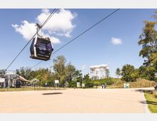 Eine Seilbahngondel schwebt über einem Park mit gepflastertem Weg, Bäumen und Rasenflächen; im Hintergrund ist eine weiße, moderne Aussichtsplattform zu sehen. Der Himmel ist klar mit vereinzelten Wolken.