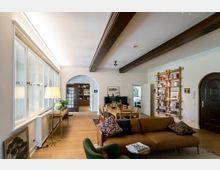 Wohnzimmer mit offener Gestaltung, großen Fenstern auf der linken Seite, einem Esstisch mit Stühlen in der Mitte und einer braunen Couch im Vordergrund. An der rechten Wand stehen ein Bücherregal und eine geschwungene Holztür, Holzbalken ziehen sich durch die Decke.