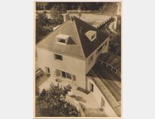 Luftaufnahme eines mehrstöckigen Hauses mit einem steilen Giebeldach, mehreren Dachfenstern und einer Terrasse. Im Vordergrund sind Bäume zu sehen, während im Hintergrund eine Straße und weitere Gebäude erkennbar sind.